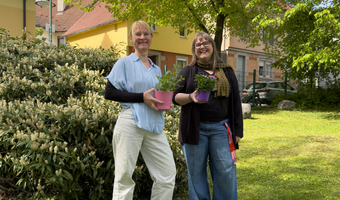 Tina Wenk links und Johanna Greef stehen im Freien vor einem Wohngebäude und halten jeweils einen Blumentopf mit Pflanzen in den Händen. Beide lächeln in die Kamera, im Hintergrund sind Sträucher und ein Baum zu sehen.