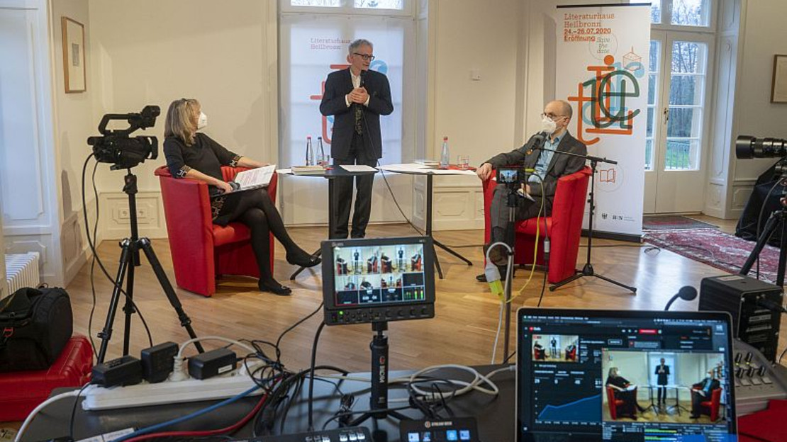 Das Bild zeigt zwei Menschen mit Corona-Schutzmasken in Sesseln im Literaturhaus aus der Anfangszeit. Im Vordergrund ist viel Videotechnik zu sehen, damals wurden Autorenlesungen nur online angeboten.