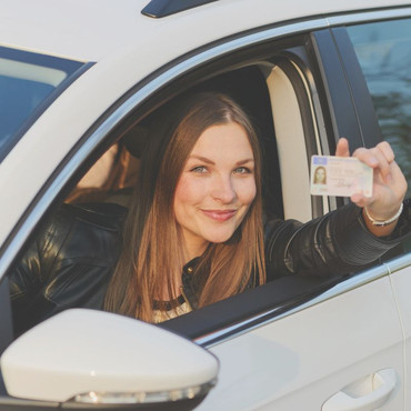 Eine Frau sitzt im Auto und lehnt sich aus dem Fahrerfenster. Sie hält einen Führerschein in die Kamera und lächelt.