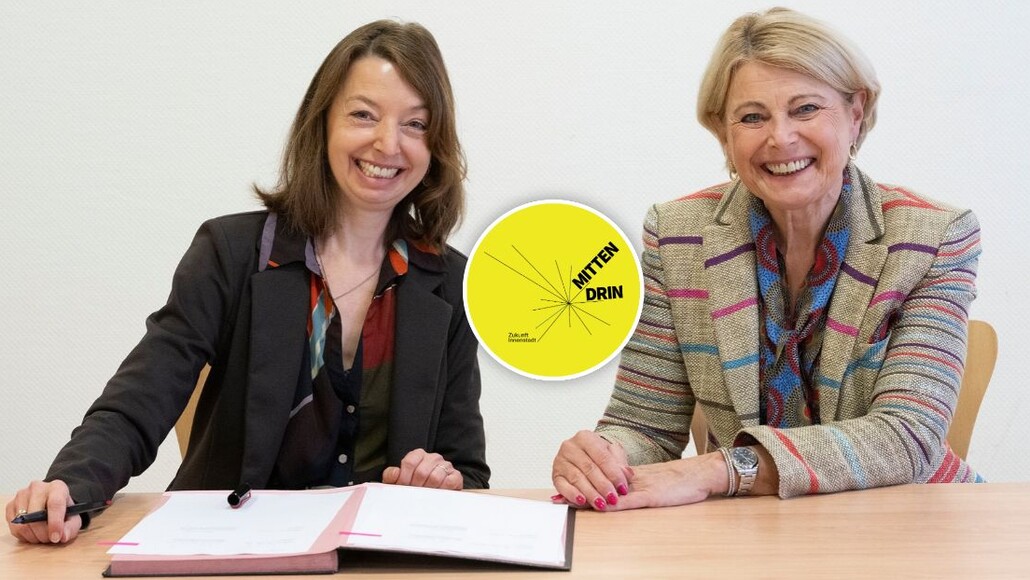 Nadine Bernecker und Agnes Christner sitzen am Tisch vor den unterzeichneten Vertragsunterlagen. Beide Frauen lächeln frlöhlich in die Kamera. In der Linken Ecke des Bildes ist das Logo "Mittendrin" für das Sofortprogramm Zukunft Innenstadt abgebildet.