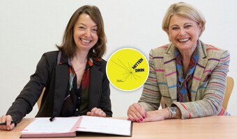 Nadine Bernecker und Agnes Christner sitzen am Tisch vor den unterzeichneten Vertragsunterlagen. Beide Frauen lächeln frlöhlich in die Kamera. In der Linken Ecke des Bildes ist das Logo "Mittendrin" für das Sofortprogramm Zukunft Innenstadt abgebildet.