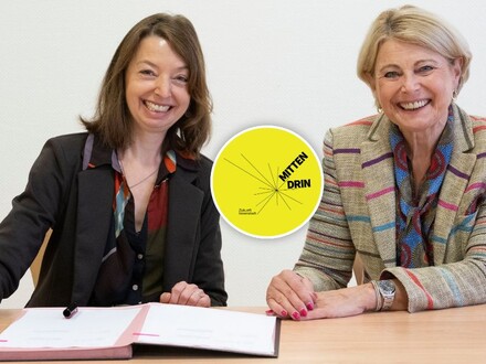 Nadine Bernecker und Agnes Christner sitzen am Tisch vor den unterzeichneten Vertragsunterlagen. Beide Frauen lächeln frlöhlich in die Kamera. In der Linken Ecke des Bildes ist das Logo "Mittendrin" für das Sofortprogramm Zukunft Innenstadt abgebildet.