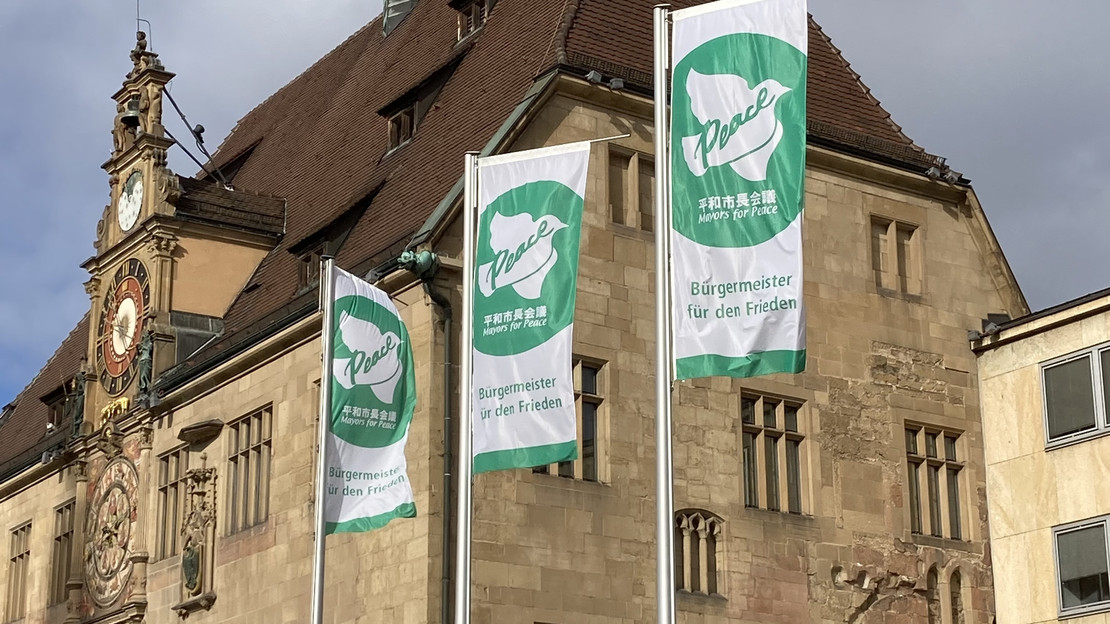 Das Bild zeigt drei wehende grüne Farben mit der weißen Friedenstaube vor dem Rathaus. Es sind Flaggen der internationalen Organisation "Mayors for Peace", übersetzt Bürgermeister für Frieden.