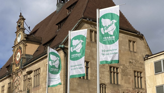 Das Bild zeigt drei wehende grüne Farben mit der weißen Friedenstaube vor dem Rathaus. Es sind Flaggen der internationalen Organisation "Mayors for Peace", übersetzt Bürgermeister für Frieden. 