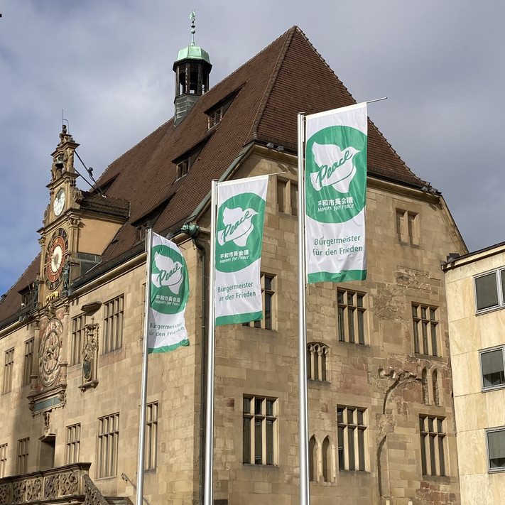 Das Bild zeigt drei wehende grüne Farben mit der weißen Friedenstaube vor dem Rathaus. Es sind Flaggen der internationalen Organisation "Mayors for Peace", übersetzt Bürgermeister für Frieden. 