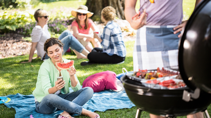 Eine Gruppe Freunde sitzt gemeinsam im Park und feiert ein Grillfest. Ein Teil der Gruppe sitzt im Hintergrund und unterhält sich. Im Vordergrund sitzt eine Frau, die in der einen Hand eine Scheibe Wassermelone und in der anderen Hand ein Handy hält, auf das sie schaut. Rechts steht ein Mann mit Schürze vor einem voll gepackten Grill und hält eine Grillzange.