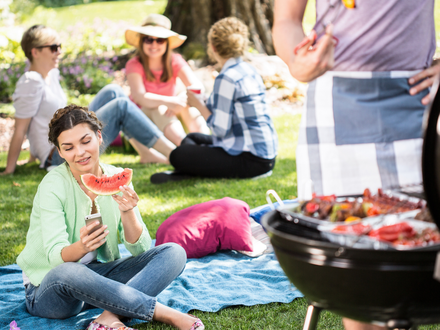 Eine Gruppe Freunde sitzt gemeinsam im Park und feiert ein Grillfest. Ein Teil der Gruppe sitzt im Hintergrund und unterhält sich. Im Vordergrund sitzt eine Frau, die in der einen Hand eine Scheibe Wassermelone und in der anderen Hand ein Handy hält, auf das sie schaut. Rechts steht ein Mann mit Schürze vor einem voll gepackten Grill und hält eine Grillzange.