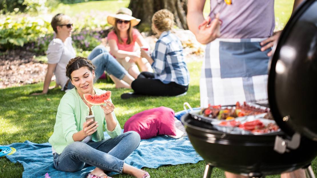 Eine Gruppe Freunde sitzt gemeinsam im Park und feiert ein Grillfest. Ein Teil der Gruppe sitzt im Hintergrund und unterhält sich. Im Vordergrund sitzt eine Frau, die in der einen Hand eine Scheibe Wassermelone und in der anderen Hand ein Handy hält, auf das sie schaut. Rechts steht ein Mann mit Schürze vor einem voll gepackten Grill und hält eine Grillzange.