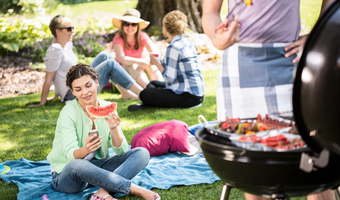 Eine Gruppe Freunde sitzt gemeinsam im Park und feiert ein Grillfest. Ein Teil der Gruppe sitzt im Hintergrund und unterhält sich. Im Vordergrund sitzt eine Frau, die in der einen Hand eine Scheibe Wassermelone und in der anderen Hand ein Handy hält, auf das sie schaut. Rechts steht ein Mann mit Schürze vor einem voll gepackten Grill und hält eine Grillzange.