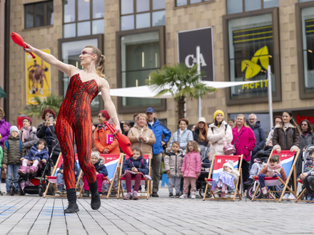 Das Bild zeigt eine Frau in rotem Kostüm auf dem Kiliansplatz, die am Kultursamstag mit Keulen jongliert und Passantinnen und Passanten unterhält. 