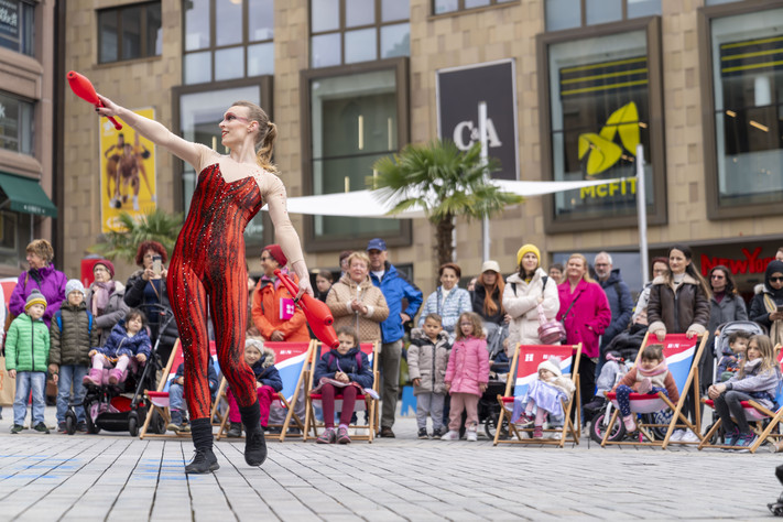Das Bild zeigt eine Frau in rotem Kostüm auf dem Kiliansplatz, die am Kultursamstag mit Keulen jongliert und Passantinnen und Passanten unterhält. 