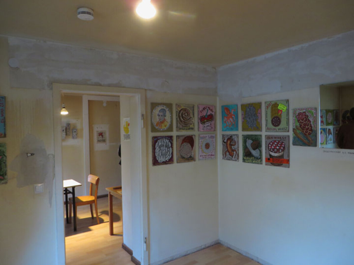 Blick in die Innenräume der Neuen Kunst: Das Bild zeigt einen kleinen Ausstellungsraum der Neuen Kunst Hagenbucher. An den weißen Wänden hängen mehrere farbintensive, quadratische Kunstwerke in einer Reihe. Durch eine offene Tür ist ein weiterer Raum mit Tisch und Stühlen sichtbar. Die Atmosphäre wirkt intim und lädt zur näheren Betrachtung der Kunst ein.
