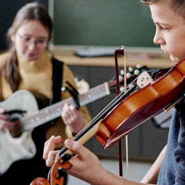 Zwei Jugendliche musizieren gemeinsam in einem Unterrichtsraum. Im Vordergrund spielt ein Junge Geige, im Hintergrund begleitet ihn ein Mädchen auf der Gitarre.