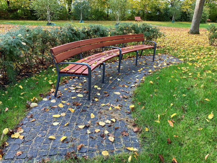 Das Bild zeigt eine gepflasterte Fläche mit einer geschwungenen Bank am Rand der Gräber-im-Park-Zone. Ausruhen oder Unterhaltung ist hier möglich. 