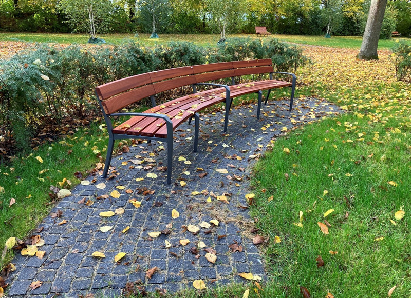 Das Bild zeigt eine gepflasterte Fläche mit einer geschwungenen Bank am Rand der Gräber-im-Park-Zone. Ausruhen oder Unterhaltung ist hier möglich. 