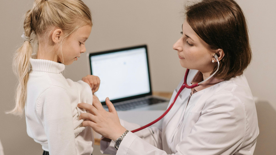 Das Bild zeigt eine Kinderärztin in weißer Arbeitskleidung, die ein junges Mädchen abhört.