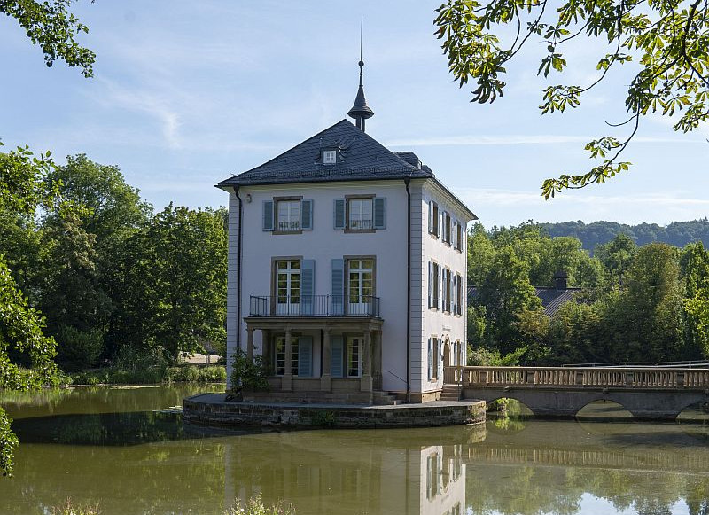 Das Bild zeigt das 450 Jahre alte Trappenseeschlösschen im Trappensee mit Zugang über eine Brücke. Das Schloss ist der Standort des Literaturhauses.