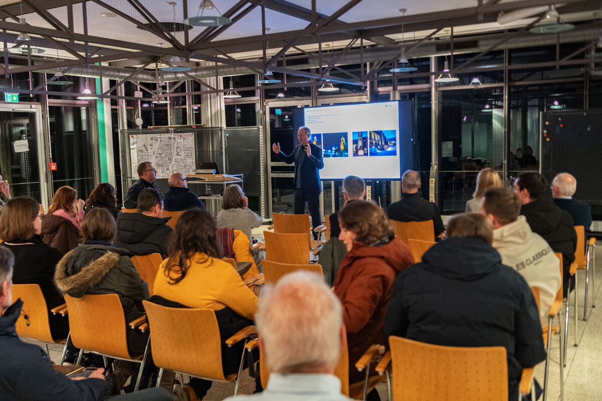Lichtplaner erklärt vor einer Powerpointpräsentation einem vollen Saal etwas zum Thema Licht