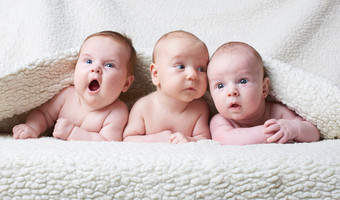 Das Bild zeigt drei Babys, die auf einer Decke liegen und sich neugierig unschauen.