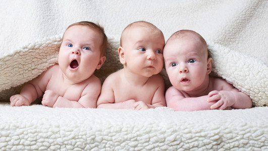 Das Bild zeigt drei Babys, die auf einer Decke liegen und sich neugierig unschauen.