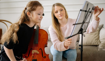 Das Bild zheigt ein Mädchen mit einem Streichinstrument, das interessiet in Notenblätter blickt. Eine Lehrerin richtet ihr die Seiten mit den Noten ein.
