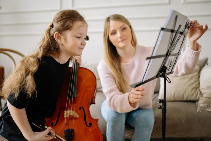 Das Bild zheigt ein Mädchen mit einem Streichinstrument, das interessiet in Notenblätter blickt. Eine Lehrerin richtet ihr die Seiten mit den Noten ein.