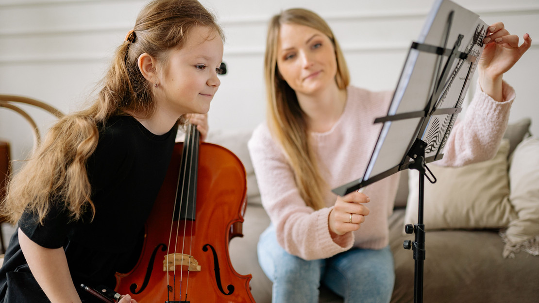 Das Bild zheigt ein Mädchen mit einem Streichinstrument, das interessiet in Notenblätter blickt. Eine Lehrerin richtet ihr die Seiten mit den Noten ein.