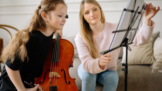 Das Bild zheigt ein Mädchen mit einem Streichinstrument, das interessiet in Notenblätter blickt. Eine Lehrerin richtet ihr die Seiten mit den Noten ein. 