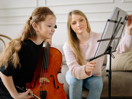 Das Bild zheigt ein Mädchen mit einem Streichinstrument, das interessiet in Notenblätter blickt. Eine Lehrerin richtet ihr die Seiten mit den Noten ein.