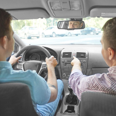 Zwei Männer sitzen in einem Auto. Der Fahrer hält das Lenkrad, der Beifahrer zeigt auf das Armaturenbrett.