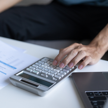 Eine Hand, die zum Taschenrechner greift, der auf einem Tisch zwischen Papieren und einem Laptop liegt.