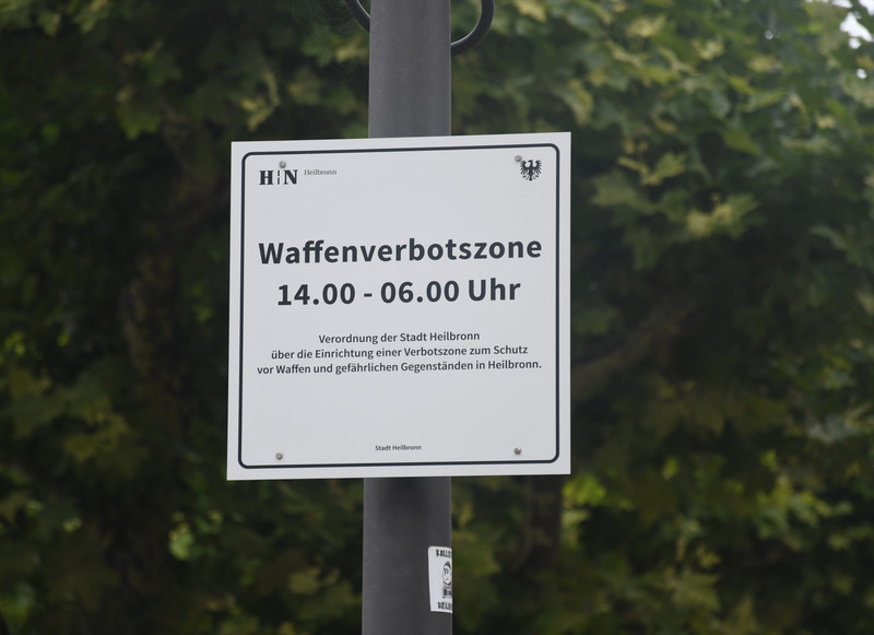 Man sieht ein Schild an einem Mast, das auf die Waffenverbotszone Innenstadt mit der Geltungszeit von 14 Uhr bis 6 Uhr morgens hinweist.