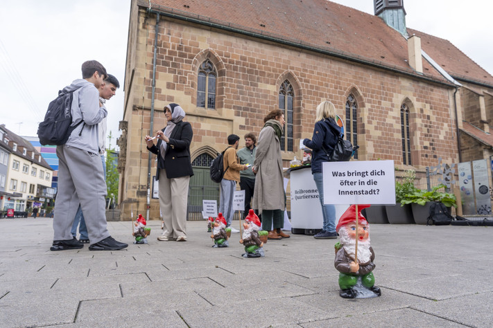 Das Bild zeigt den Infostand der Bürgerbeteilgung in der Sülmerstraße. Dort kommen Mitarbeitende der Stadt mit Bürgerinnen und Bürgern zur Entwicklung der Innenstadt und des neuen Quartiersbüros ins Gespräch. Zwergfiguren mit Fragen zur Innenstadt machen auf den Stand aufmerksam.