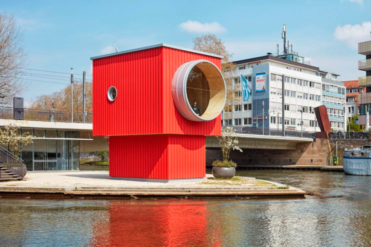 One Man House von Thomas Schütte: Das Bild zeigt das One Man House in Heilbronn, ein auffälliges, würfelförmiges Gebäude in kräftigem Rot. Es steht auf einer Plattform direkt im Wasser. An der Vorderseite befindet sich eine große runde Öffnung in heller Farbe, die wie ein Fenster oder Tunnel wirkt. Im Hintergrund sind moderne Gebäude, eine Brücke und städtische Elemente zu sehen. Die Wasseroberfläche spiegelt das Gebäude und verstärkt die besondere Wirkung dieses Kunstobjekts im öffentlichen Raum.