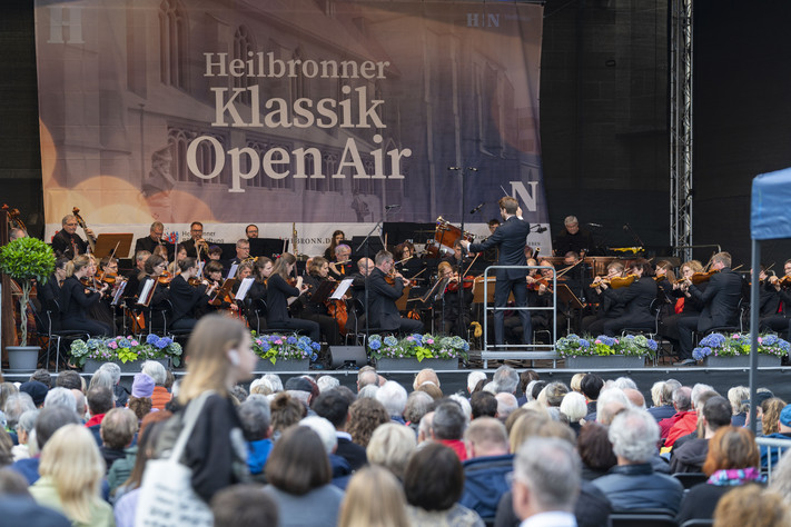 Das Bild zeigt eine große Bühne auf dem Kiliansplatz mit einem Orchester. Auf dem Platz sitzen viele Zuschauerinnen und Zuschauer und lauschen den Klängen.