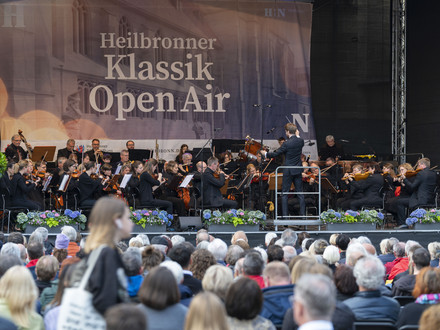 Das Bild zeigt eine große Bühne auf dem Kiliansplatz mit einem Orchester. Auf dem Platz sitzen viele Zuschauerinnen und Zuschauer und lauschen den Klängen.