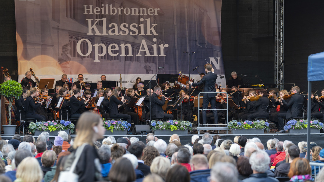 Das Bild zeigt eine große Bühne auf dem Kiliansplatz mit einem Orchester. Auf dem Platz sitzen viele Zuschauerinnen und Zuschauer und lauschen den Klängen.