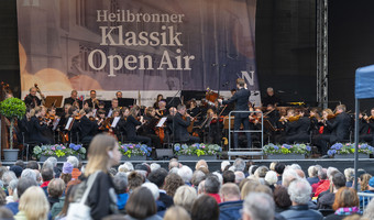 Das Bild zeigt eine große Bühne auf dem Kiliansplatz mit einem Orchester. Auf dem Platz sitzen viele Zuschauerinnen und Zuschauer und lauschen den Klängen.