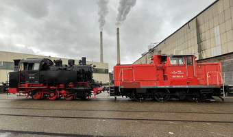 Das Bild zeigt eine rote Rangierlok, die auf den Gleiser der Hafenbahn eine historsiche Dampflok zum Verladen auf ein Schiff zieht.