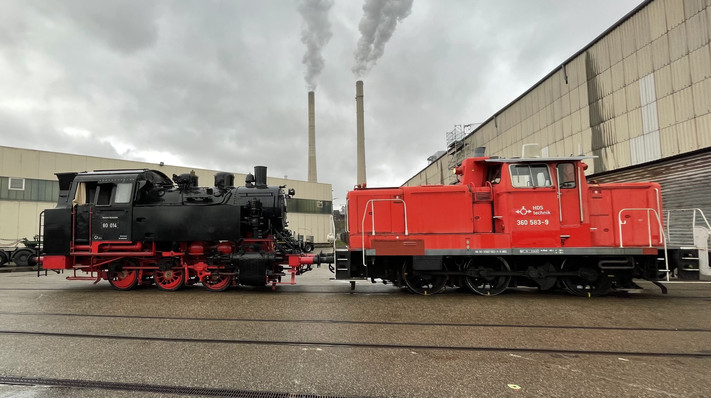 Das Bild zeigt eine rote Rangierlok, die auf den Gleiser der Hafenbahn eine historsiche Dampflok zum Verladen auf ein Schiff zieht.