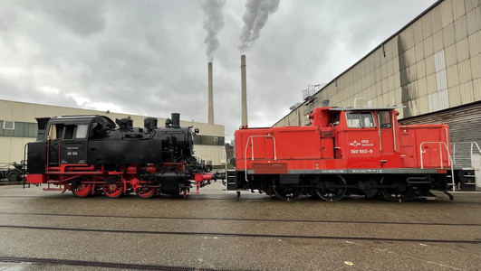 Das Bild zeigt eine rote Rangierlok, die auf den Gleiser der Hafenbahn eine historsiche Dampflok zum Verladen auf ein Schiff zieht. 