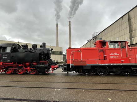 Das Bild zeigt eine rote Rangierlok, die auf den Gleiser der Hafenbahn eine historsiche Dampflok zum Verladen auf ein Schiff zieht.