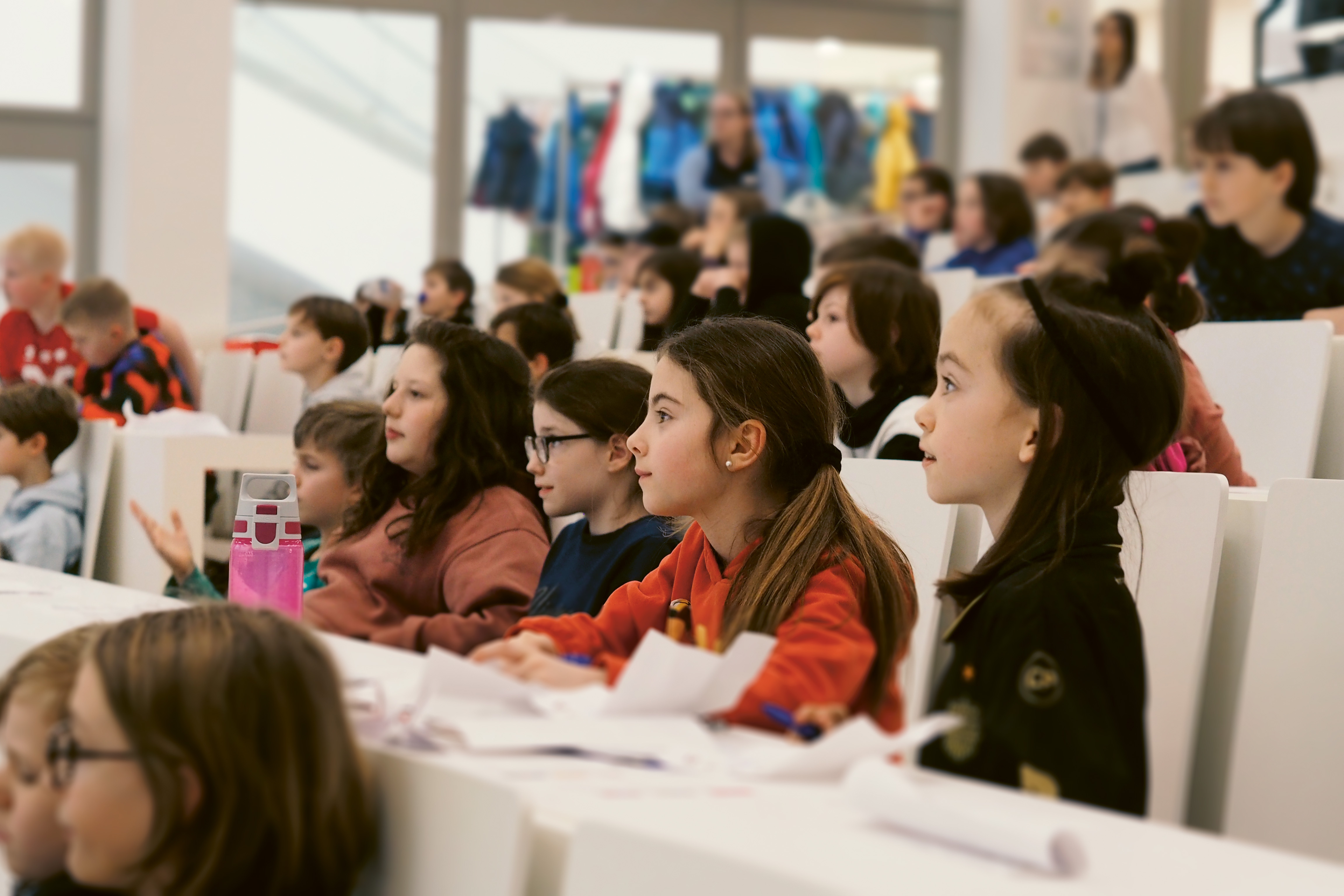 Man sieht ein Bild der aim Kinderuni. Viele Kinder sitzen aufmerksam in einem Hörsaal und lauschen den Ausführungen der Lehrperson.
