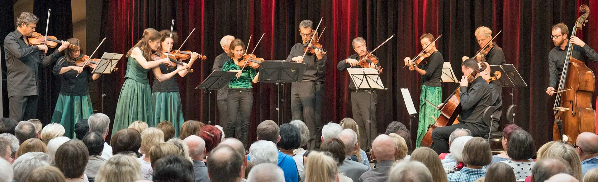Das Bild zeigt ein Ensemble von Musikerinnen und Musikern auf einer Bühne mit rotem Vorhang im Hintergrund. Sie spielen Streichinstrumente wie Violinen, Bratschen und Kontrabass. Vor der Bühne sitzt ein Publikum, das dem Konzert aufmerksam zuhört. Die Szene vermittelt eine klassische Konzertatmosphäre und kulturelles Leben.