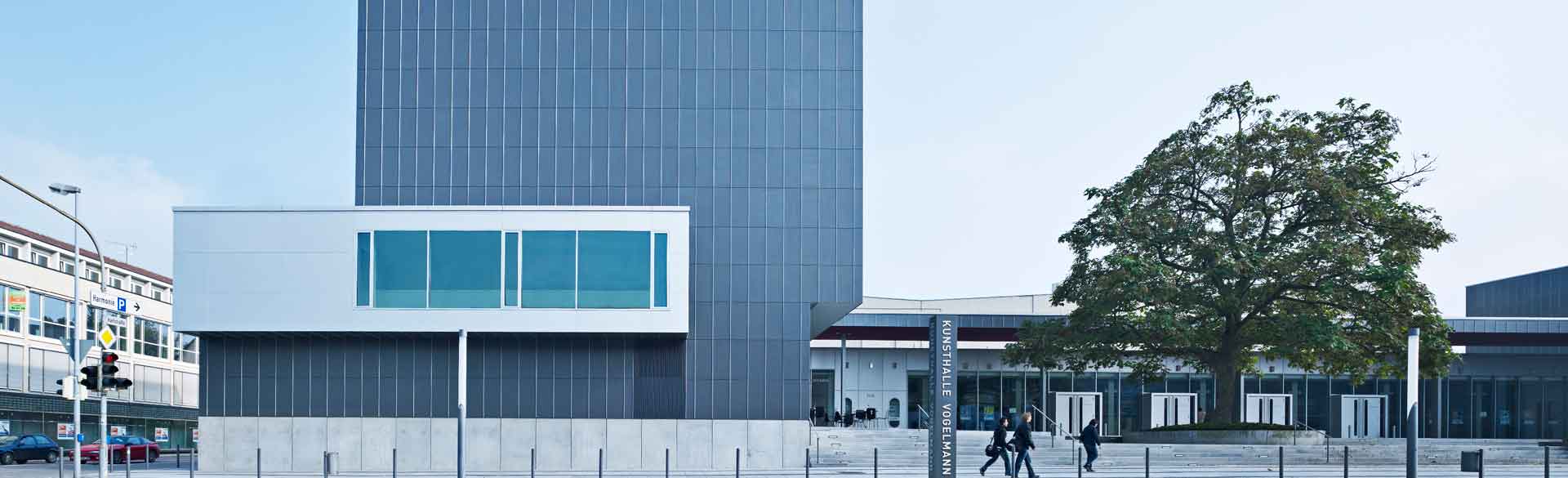 Das Bild zeigt die Kunsthalle Vogelmann in Heilbronn von außen. Das Gebäude ist modern gestaltet mit klaren Linien, großen glatten Fassaden und einem hervorgehobenen, rechteckigen Anbau. Vor dem Gebäude verläuft ein offener Platz, auf dem sich einige Menschen bewegen. Rechts steht ein großer Baum, der einen Kontrast zur sachlichen Architektur bildet. Die Szene vermittelt einen modernen, kulturellen Ort im städtischen Raum.