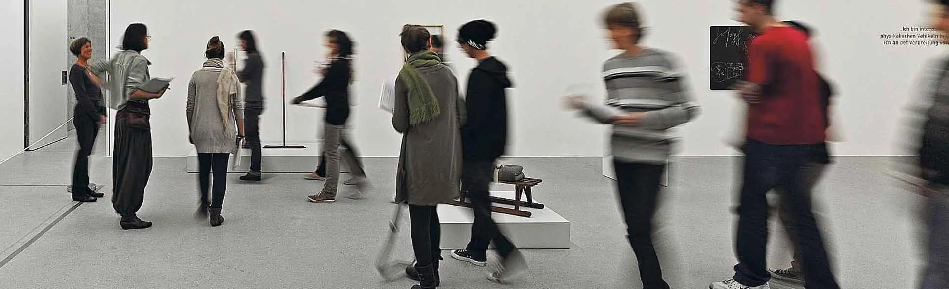 Headerbild: Das Bild zeigt den Innenraum der Kunsthalle Vogelmann mit mehreren Personen, die sich durch eine Ausstellung bewegen. Einige bleiben stehen und betrachten Kunstwerke an den Wänden, während andere durch den Raum gehen. Die Umgebung ist hell und minimalistisch gestaltet, wodurch die Kunstwerke und die Bewegung der Besucherinnen und Besucher im Mittelpunkt stehen.