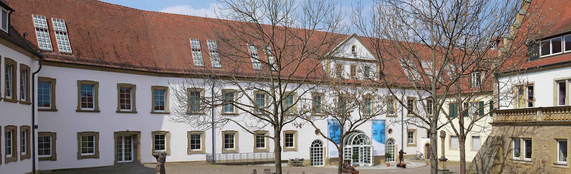 Das Bild zeigt das Museum im Deutschhof in Heilbronn. Mehrere historische Gebäude mit hellen Fassaden und roten Dächern sind um einen offenen Platz angeordnet. Vor den Gebäuden stehen mehrere Bäume ohne Blätter. Der Eingangsbereich ist zentral gelegen und gut erkennbar. Die Szene vermittelt den historischen Charakter des Museumsensembles und seine ruhige, offene Umgebung.