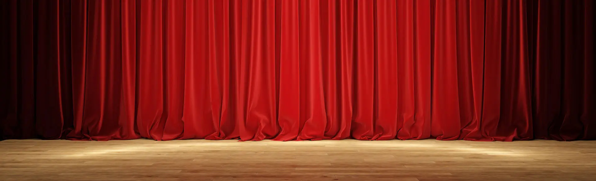 Ein Headerbild: Das Bild zeigt eine Theaterbühne mit einem vollständig geschlossenen, roten Vorhang. Der Vorhang ist in Falten gelegt und reicht bis zum Boden. Davor ist ein leerer Bühnenboden aus Holz zu sehen. Die Szene wirkt ruhig und lässt auf eine bevorstehende Aufführung oder eine Pause schließen.