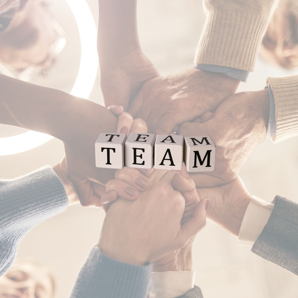 Das Bild zeigt mehrere Hände, die sich in der Mitte übereinanderlegen. Auf einigen Fingern sind kleine Buchstabenwürfel verteilt, die zusammen das Wort Team bilden. Die Szene steht für Zusammenhalt, Kooperation und gemeinsames Arbeiten in einer Gruppe.