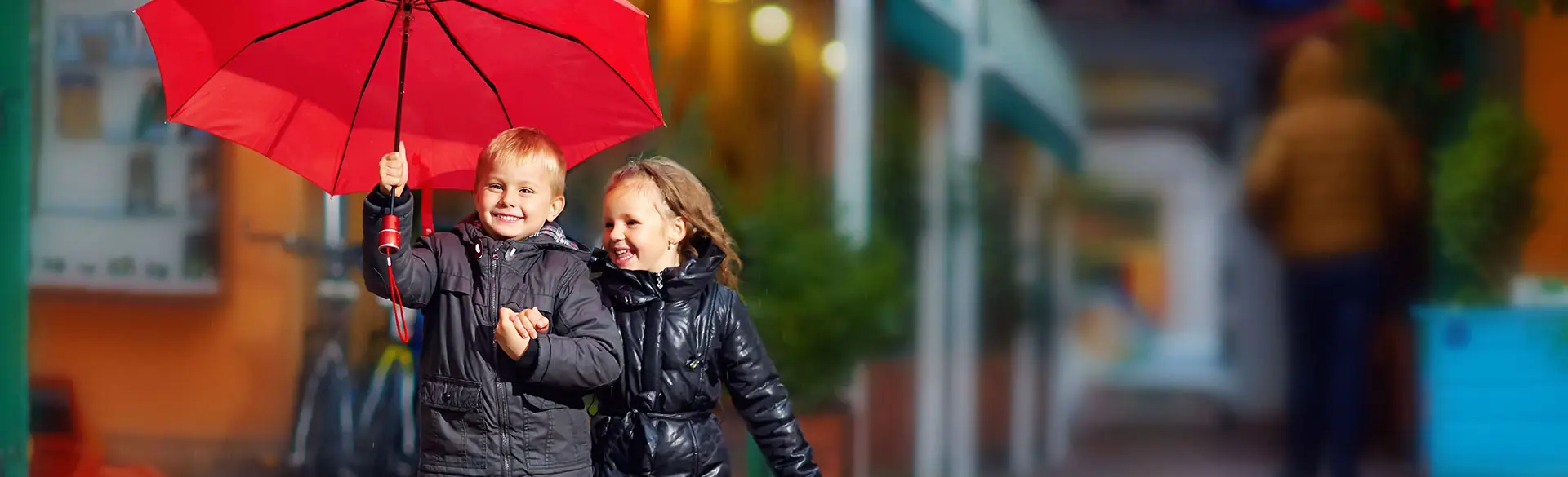 Ein Headerbild: Das Bild zeigt zwei Kinder, die nebeneinander auf einer Straße unterwegs sind. Ein Kind hält einen roten Regenschirm über beide, während sie sich an den Händen halten und lachen. Beide tragen Jacken und wirken gut geschützt vor dem Wetter. Im Hintergrund ist eine leicht unscharfe Stadtszene zu erkennen. Die Darstellung vermittelt Geborgenheit, Freude und sicheres Unterwegssein im Alltag.